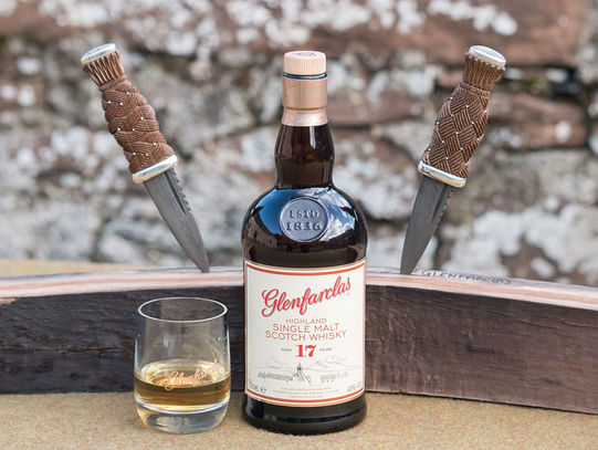 A pair of traditional Scottish Sgian Dubhs with carved handles made from a Glenfarclas whisky barrel stave. Sgian Dubhs are on a stave from a whisky barrel behind a bottle of Glenfarclas whisky and a glass with whisky in.