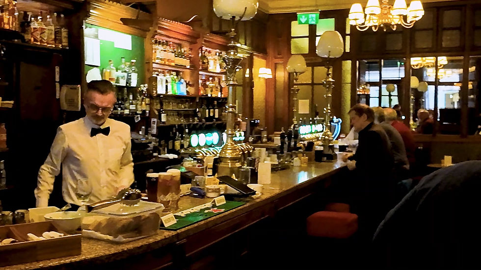 Crowded restaurant with people dining at long tables, warm lighting, and a cozy atmosphere. Glasses and bottles are visible on tables.