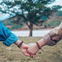 Caucasian women holding hands in front of a tree and a body of water.