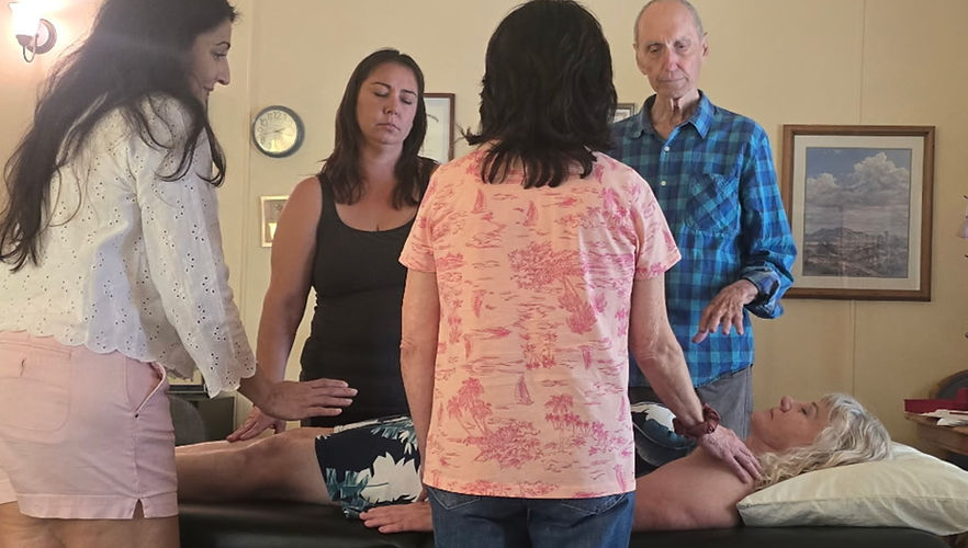 Glen Larsen and an assortment of female students with their hands above or on a middle-aged woman who is prone on a massage table.