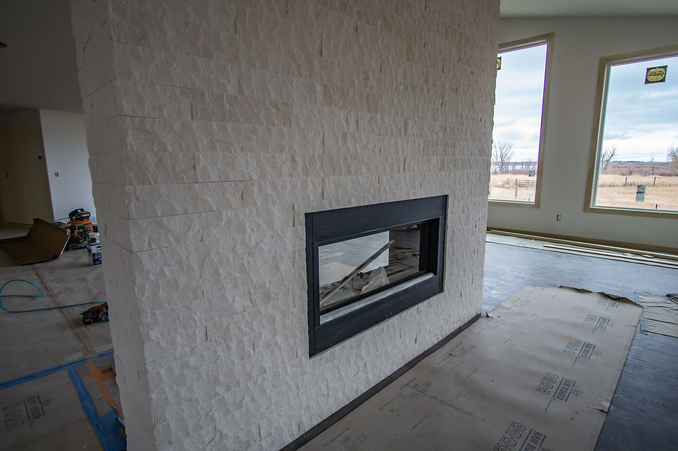 Modern living room with a white stone fireplace and large windows overlooking a field. Unfinished floor with construction materials.