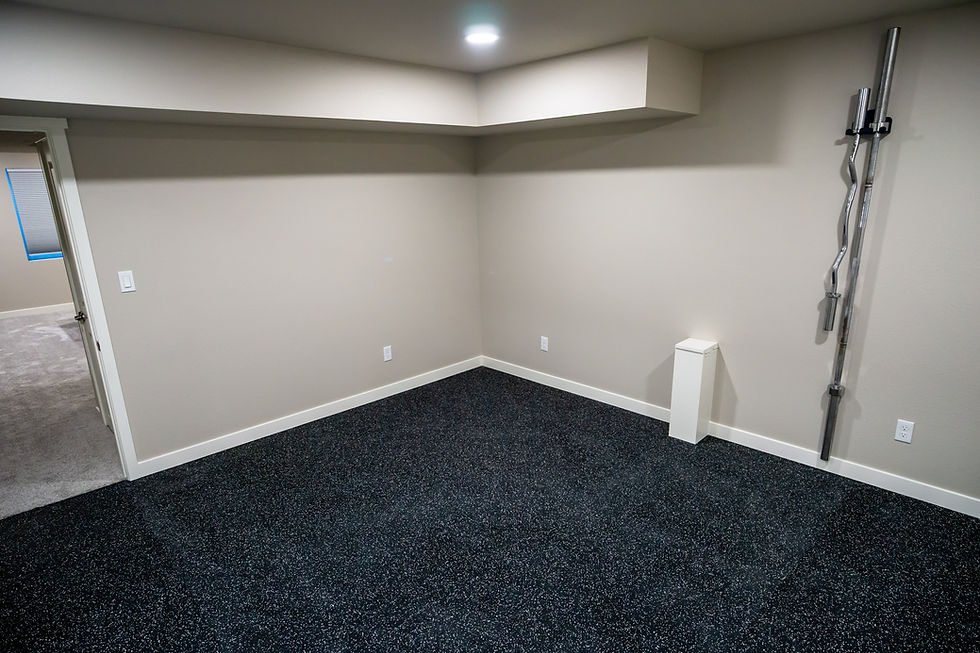 Workout room with speckled dark rubber floor, beige walls, and a mounted barbell on the wall. Open doorway leads to another room. Bright lighting.