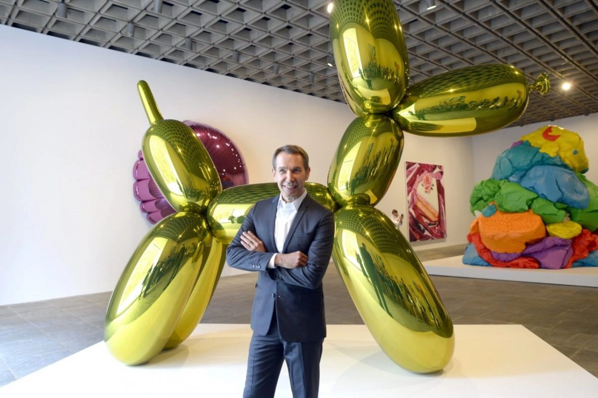 A photo of a gold-colored, balloon dog sculpture by Jeff Koons in an art gallery, surrounded by other artworks and a person.