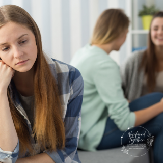 An adolescent girl sits looking towards the camera forlornly as 2 of her peers sit behind her laughing and talking together.