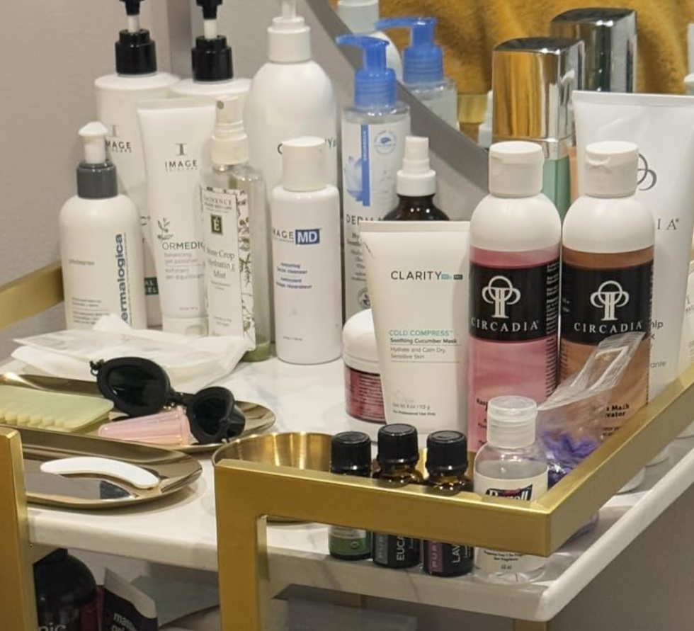 A collection of skincare products, bottles, and jars are neatly arranged on a gold-trimmed counter. Includes a small mirror and sunglasses.