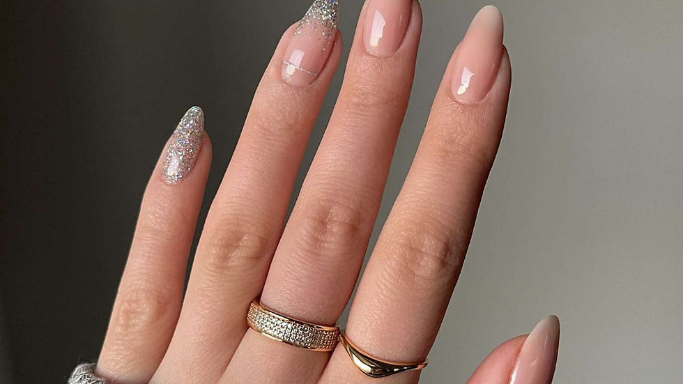 Hand with nude and glitter manicure, wearing gold rings. Gray sweater sleeve visible. Neutral background, showcasing elegant nails.