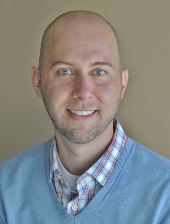 Smiling bald man in a blue sweater and plaid shirt.