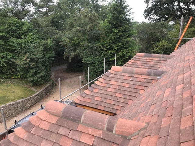 Overhead view of a newly tiled pitched roof with red clay tiles featuring carefully aligned rows and curved ridge tiles. Scaffolding is visible around the roof, with a stone wall, pathway, and lush green trees in the background.