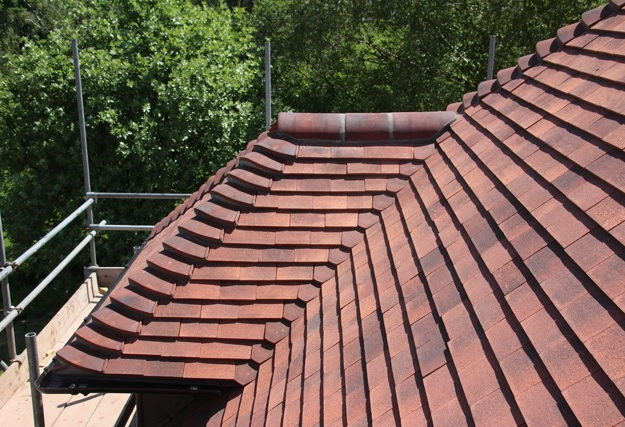 View of a newly installed pitched roof with red clay tiles, showcasing neat and uniform tile arrangement. Scaffolding is visible on the left side, with green trees in the background.