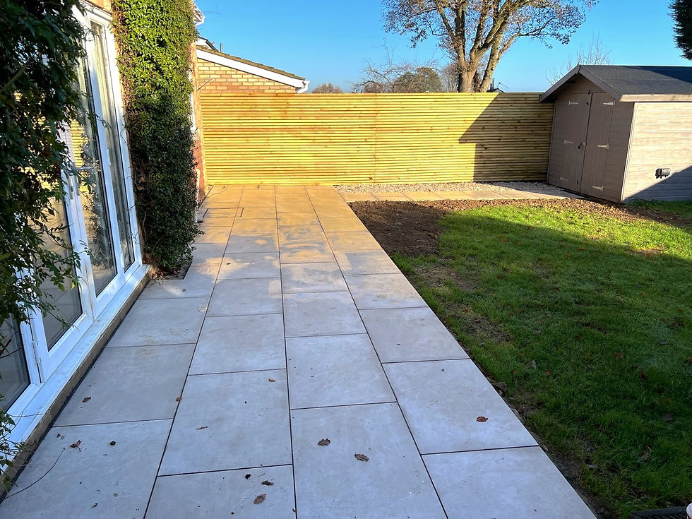Newly installed patio with large rectangular tiles in a clean, light gray finish by Mike 360 Groundworks and Landscaping, bordered by green grass on the left and leading up to a set of white double doors on the right.