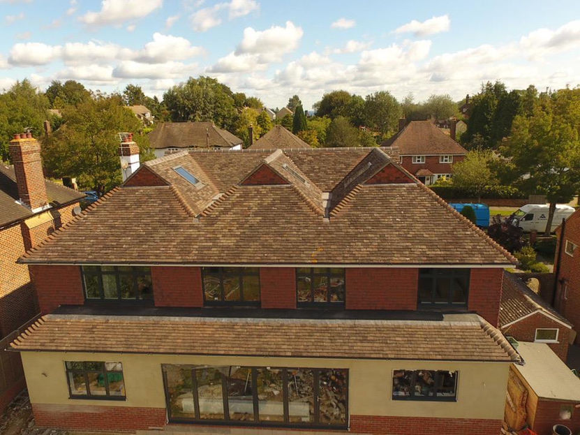 Aerial view of a large two-story house with a newly installed pitched roof featuring brown clay tiles. The roof includes dormer windows and skylights. The surrounding area has other houses, trees, and a clear sky with scattered clouds.