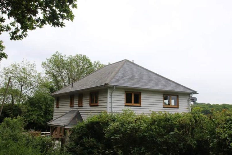 A two-story house with a light grey, pitched hip roof, surrounded by lush greenery. The house features white horizontal siding and wooden-framed windows, adding a rustic charm. The roof appears to be made of slate tiles, contributing to the home's classic and sturdy appearance. The surrounding vegetation includes various shrubs and trees, with a backdrop of more dense forest, indicating a serene and secluded location.