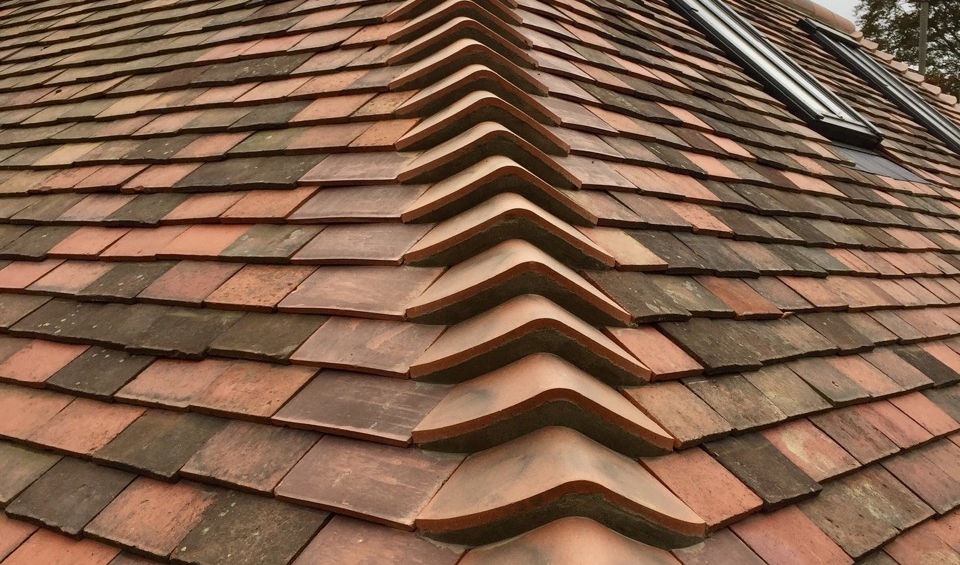Angled view of a pitched roof with red and brown shingles and a neatly aligned ridgeline. The roof features a skylight window, and the photo highlights the craftsmanship and quality of the roofing installation against a cloudy sky backdrop.