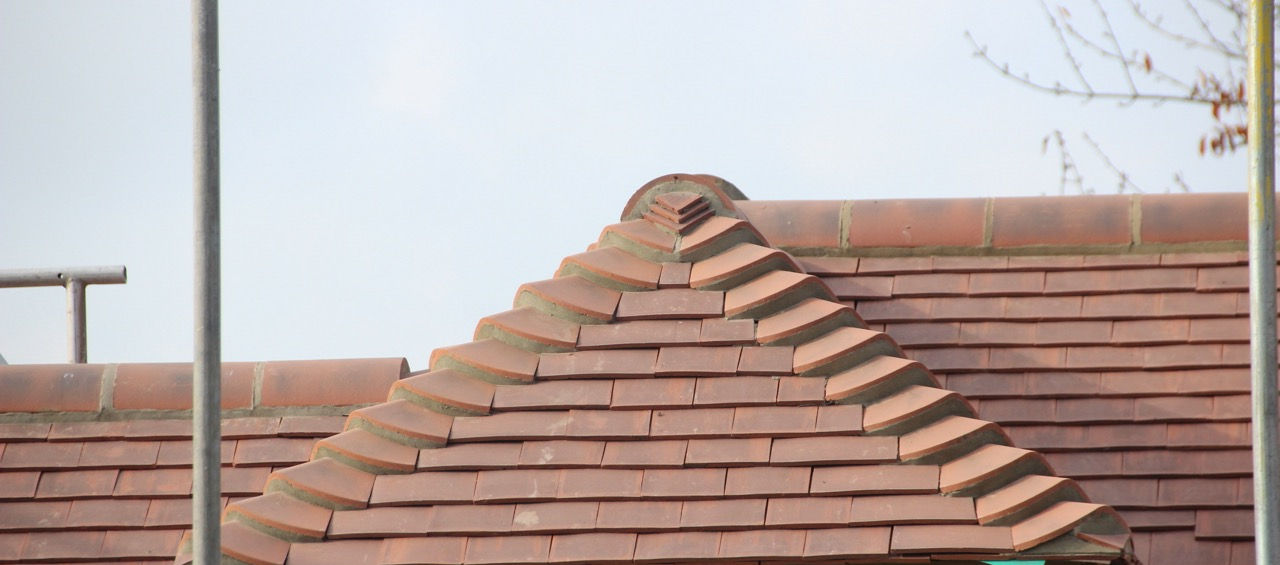A close-up view of a newly constructed pitched roof with red clay tiles. The roof's ridge tiles are prominently featured, neatly arranged in a stepped pattern, showcasing detailed craftsmanship. The eaves and underlayment are visible, with a green membrane marked "GREEN SIDE UP LAP LINE 150mm" partially exposed. Scaffolding poles are present on either side, indicating ongoing construction work. The background shows a clear sky and some branches of a nearby tree.
