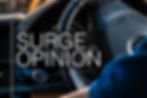 Close-up of a car interior featuring a steering wheel and dashboard. Text overlay reads "SURGE OPINION" in bold white letters.