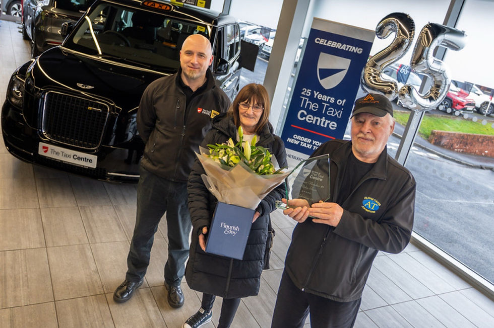 Three people stand in a The Taxi Centre showroom, one holding flowers, another holding an award. "25 Years of The Taxi Centre" balloons and sign in background.