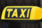Yellow taxi sign with bold black "TAXI" text, set against a blurred dark background. Bright and noticeable.