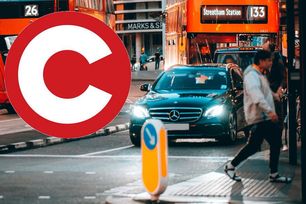 Urban street with busy traffic, red buses, and pedestrians. A large red "C" sign is in focus. The mood is bustling and active.