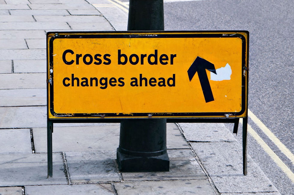 Yellow street sign on a sidewalk reads "Cross border changes ahead" with a black arrow. Road visible, suggesting a transition in traffic.