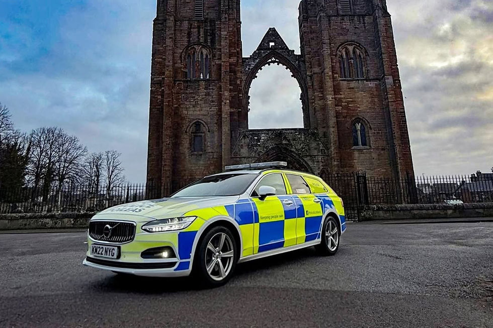 Police car with blue and yellow checks parked in front of a large, weathered stone building and archway. Overcast sky, somber mood.
