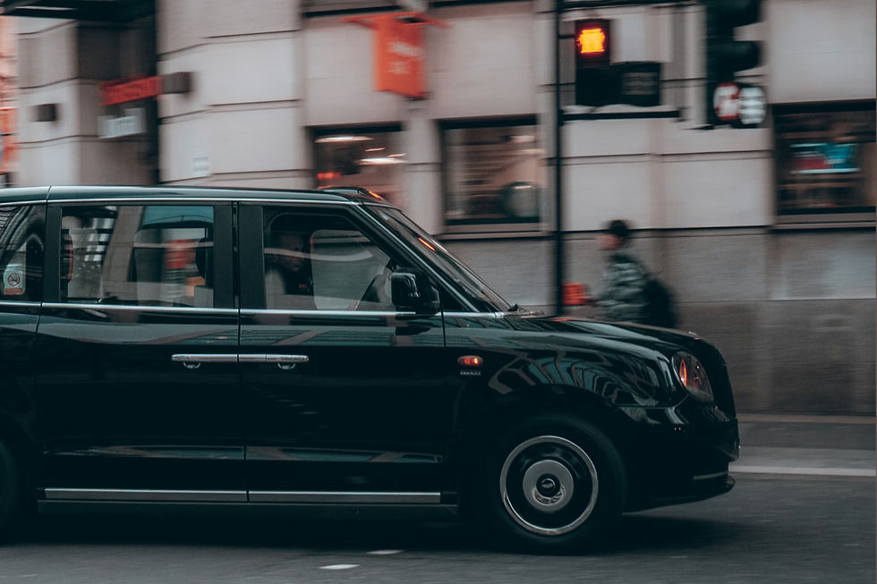 Black taxi drives past a city street with blurred pedestrian in the background. Red traffic light visible, urban atmosphere captured.