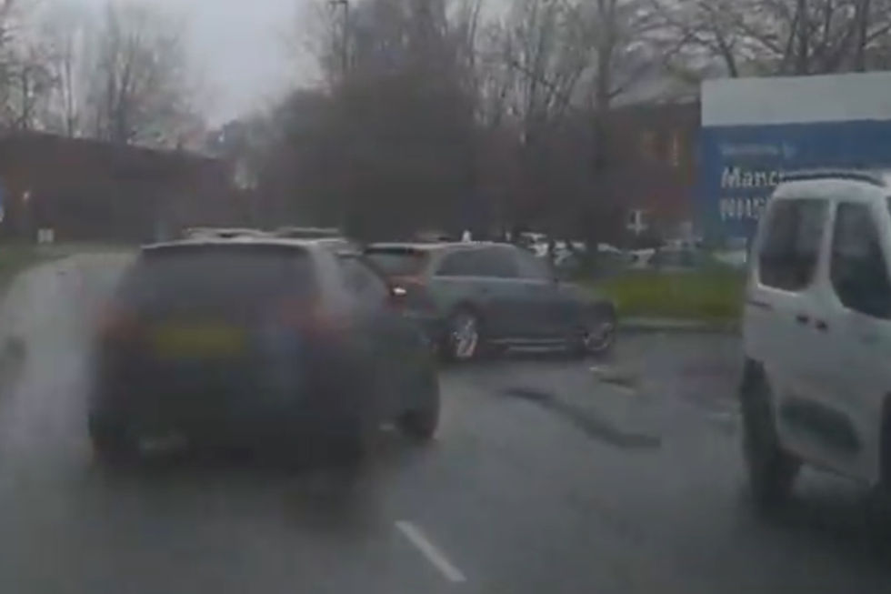 Multiple cars on a wet road near a blue "Welcome to Manchester" sign. Overcast sky, bare trees in the background, creating a gloomy mood.