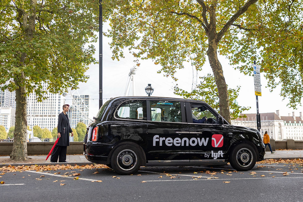 Black cab with "Freenow by Lyft" logo parked under trees with autumn leaves. Person holding red umbrella walks nearby, cityscape in background.