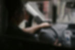 Driver's hands on steering wheel inside a car, raindrops on window, blurred cityscape outside. Dashboard controls visible, subdued mood.