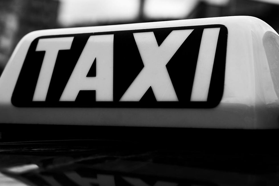 Close-up of a black and white taxi sign, with bold "TAXI" text, set against a blurred urban background. Mood is straightforward and functional.