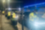 Police officers in yellow jackets conduct a nighttime traffic check beside a row of parked cars. Blue Christmas lights illuminate the background.