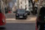 Black London taxi drives down a city street with red buses and pedestrians, under bare trees and pale buildings.