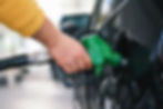 Hand in a yellow sleeve refuels a black car using a green gas nozzle. Close-up shot, outdoor setting, focus on the nozzle and hand.