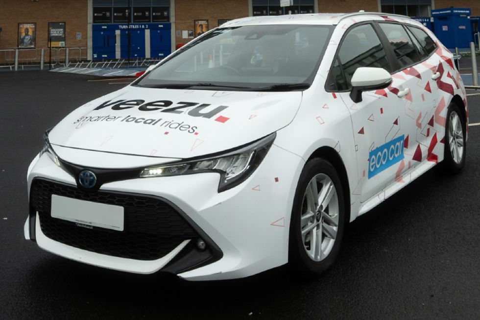 White car with red triangle patterns and "Veezu, smarter local rides" printed. "Eco car" text on side. Parked on wet asphalt.