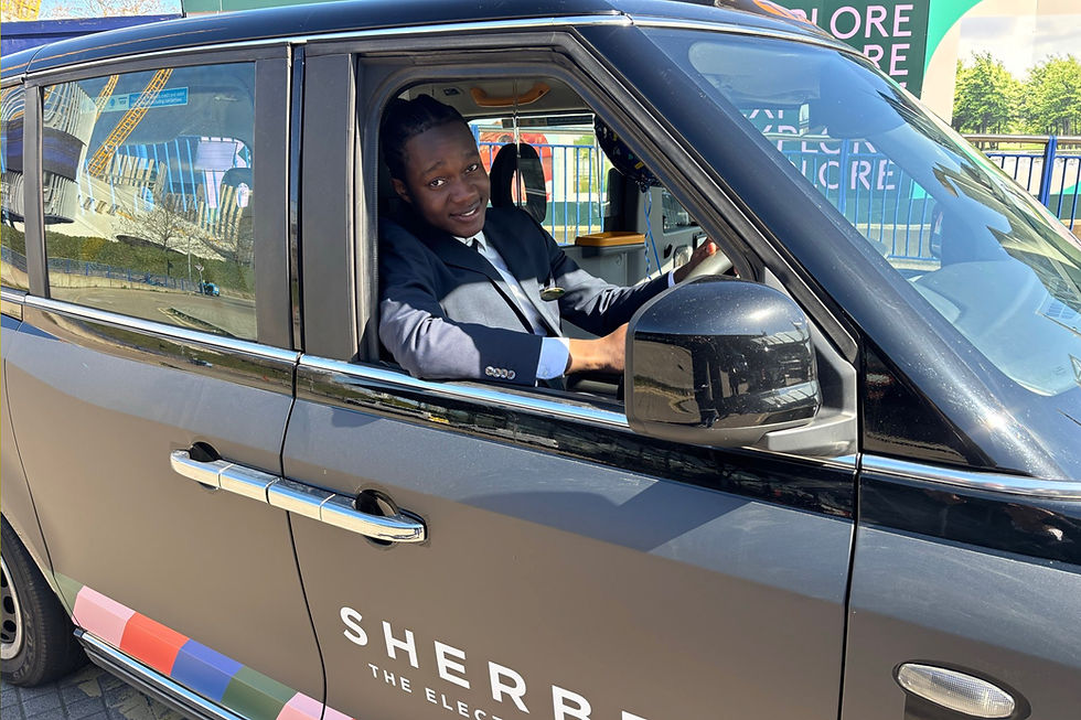 Newly qualified London taxi driver in a suit smiles inside a black London taxi with "Sherbet" on the side. Reflections of modern buildings seen on the car windows.