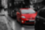 Red car in city traffic, surrounded by a black bus and other vehicles on a wet street. Monochrome background adds contrast.