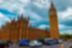 Big Ben and the Houses of Parliament under a cloudy sky. Cars and taxis on the road, with people walking by. Vibrant colors and dynamic scene.