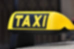Yellow taxi sign with bold black "TAXI" text, mounted on a vehicle roof. The background is blurred, suggesting an urban street setting.
