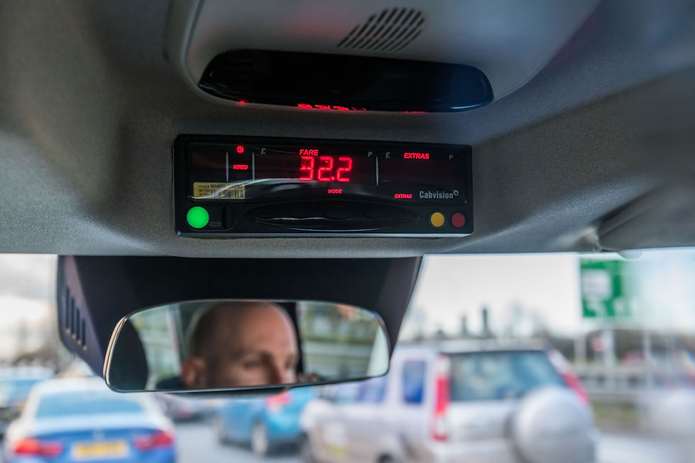 Taxi meter showing fare of £32.2, with a reflection of a bald man in the rearview mirror. Background shows cars and a blurry road sign.