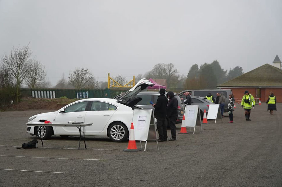 People in a car park near private hire vehicles and traffic cones, some wearing high-vis jackets. Overcast sky, bare trees in the background.