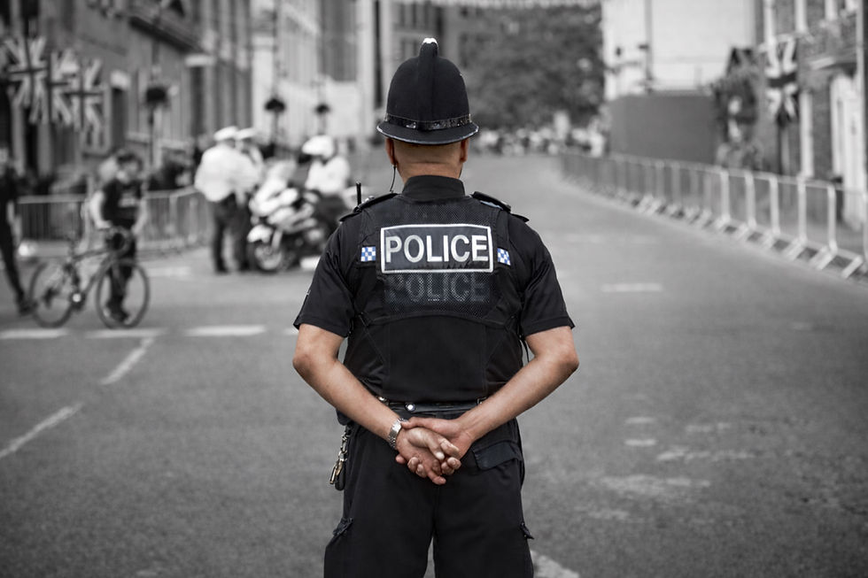 Police officer stands with hands clasped, facing street. "POLICE" visible on uniform. Blurred background with cyclists and flags. Calm scene.