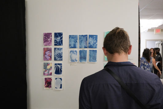 A man stands in front of an installation of purple, green and blue cyanotype pieces by Erin Grey at ARCHIVES first exhibition.