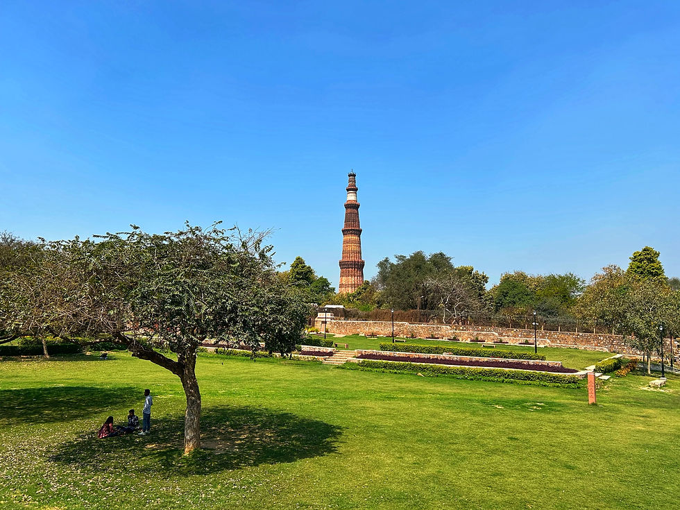 Qutub Minar stands in a sunny park with green grass and trees. People sit in the shade, enjoying a clear blue sky. Relaxed atmosphere.