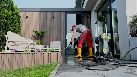 Person in red and gray clothing pressure washes a modern patio. Bright yellow boots and green plants on a wooden deck add color.