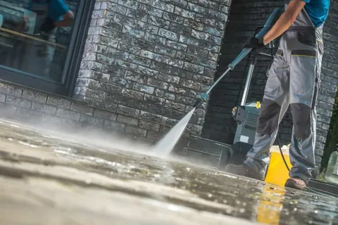 Person power-washing a wet, stone pavement in front of a brick wall. Wearing gray work clothes, using a blue hose, and standing by a yellow bucket.
