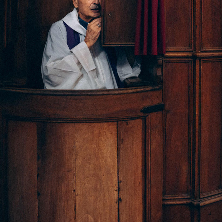 A priest in liturgical vestments standing inside a wooden confessional, looking thoughtfully toward the side.