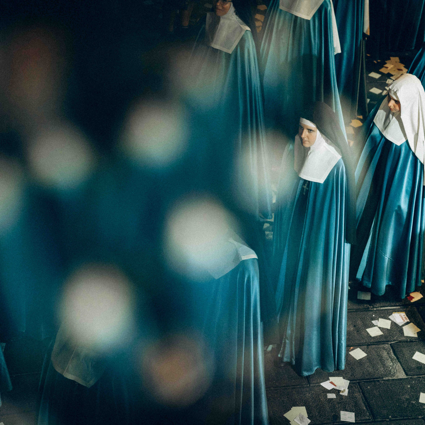 A procession of nuns in teal and white cloaks walking through a dimly lit outdoor space with petals on the floor.