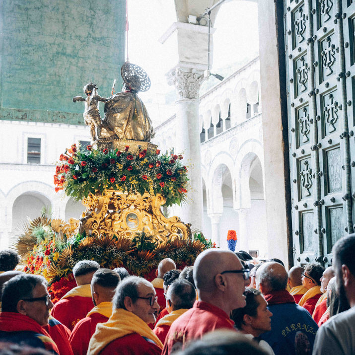 A large crowd gathered in front of church doors watching a decorated statue platform being carried outside.