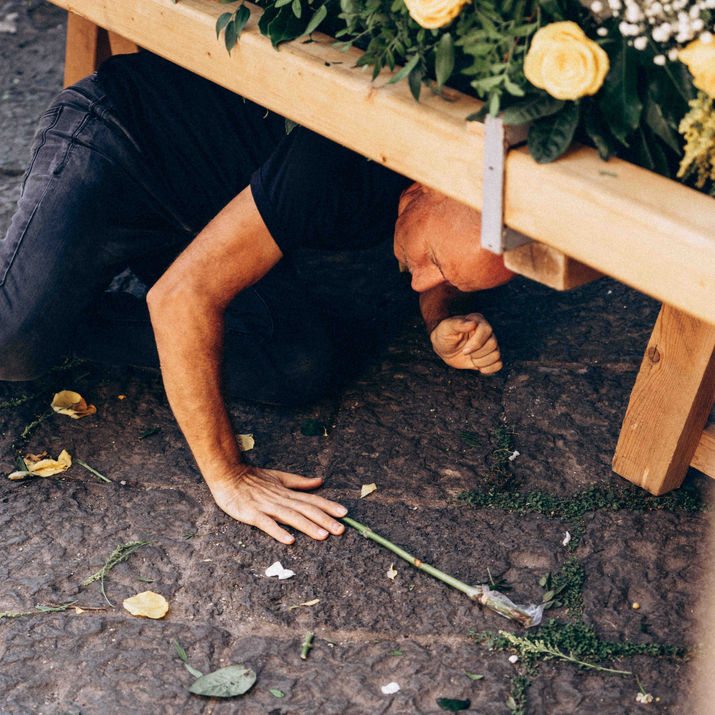 A man crawling underneath a wooden structure covered in yellow flowers, picking up a flower stem from the paved ground.