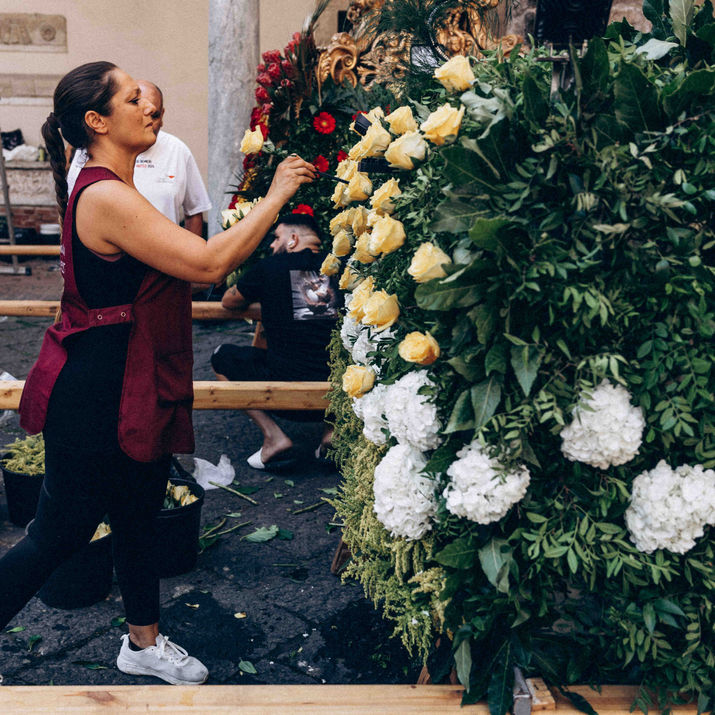 A woman arranging yellow and white flowers on a large wooden structure covered in greenery during event preparations outdoors.