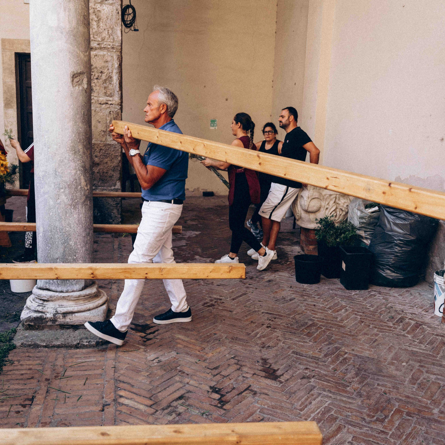 A man carrying a long wooden beam through a historic courtyard, with other people assisting in the background.